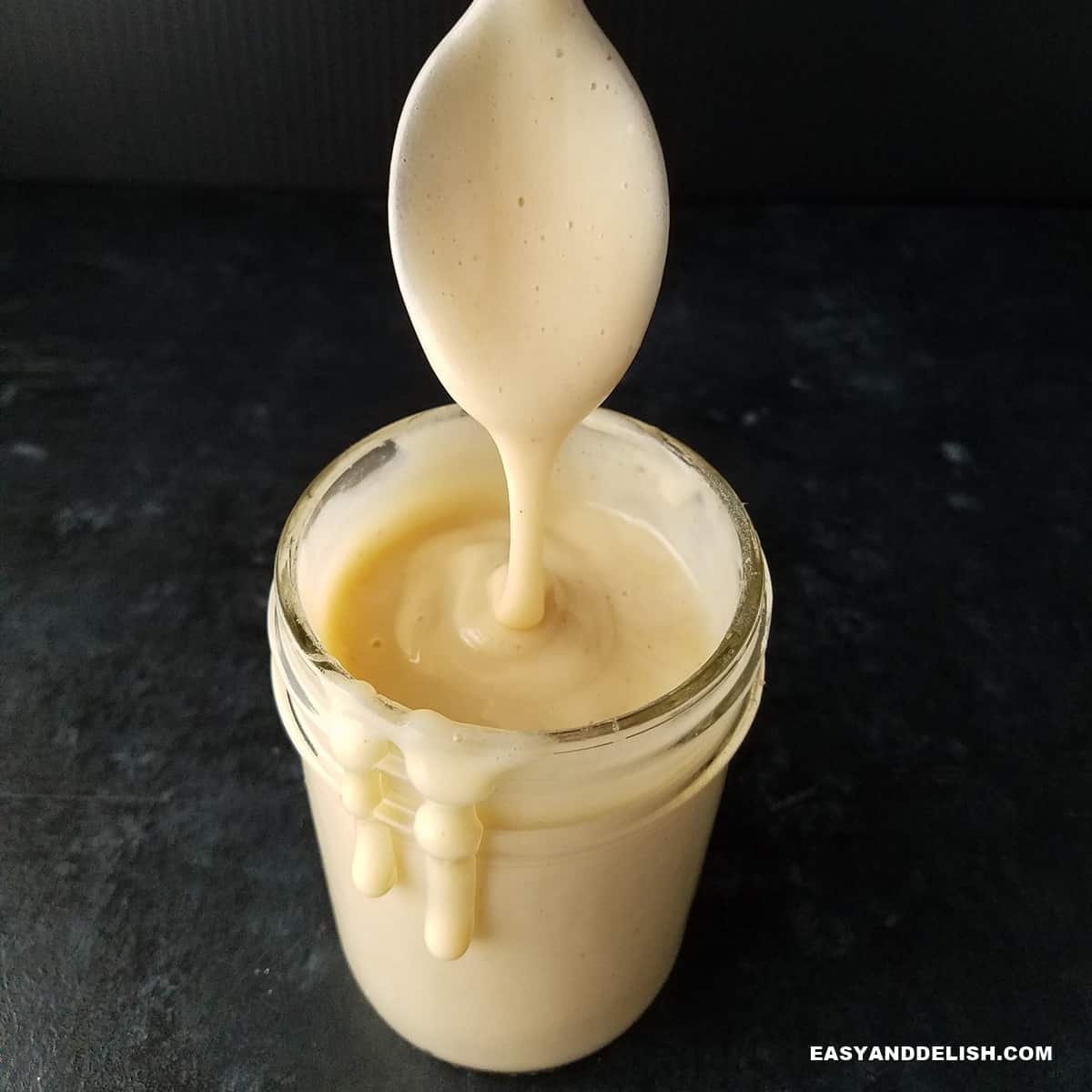 close up of a jar of sugar-free keto condensed milk dripping on the sides with a spoon held on top.