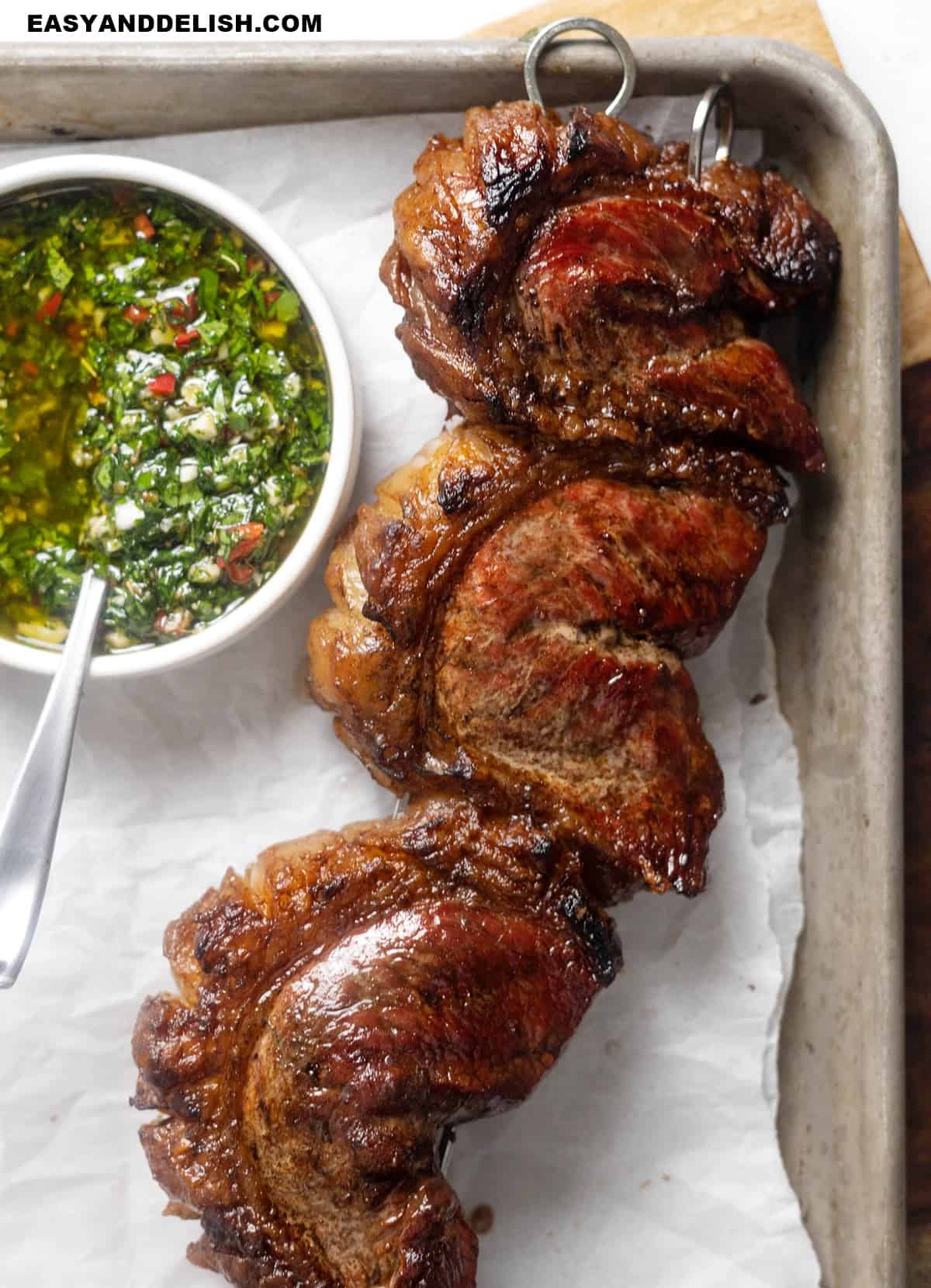 close up of a skewer of grilled picanha steaks with a bowl of chimichurri sauce on the side.