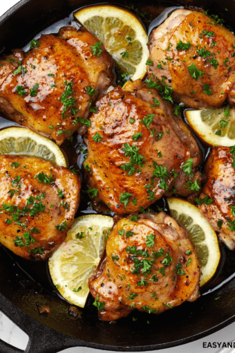 close up image showing a pan with honey butter chicken with chopped parsley and lemon wedges.