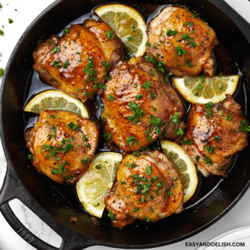 close up image showing a pan with honey butter chicken with chopped parsley and lemon wedges.