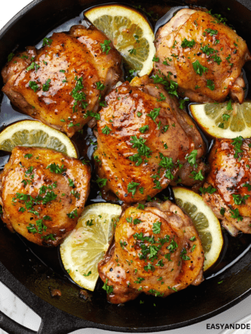 close up image showing a pan with honey butter chicken with chopped parsley and lemon wedges.