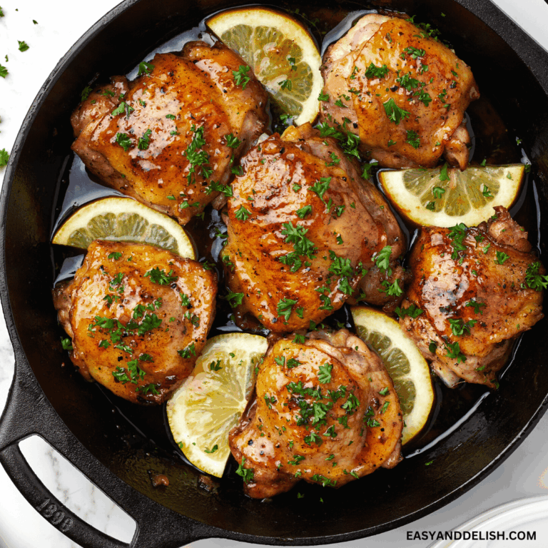 close up image showing a pan with honey butter chicken with chopped parsley and lemon wedges.