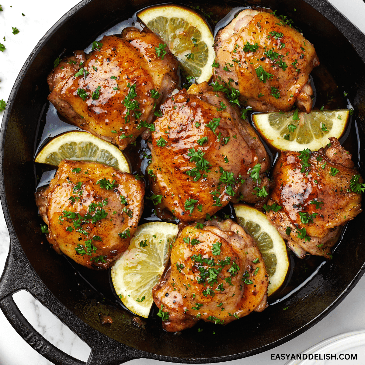 close up image showing a pan with honey butter chicken with chopped parsley and lemon wedges.