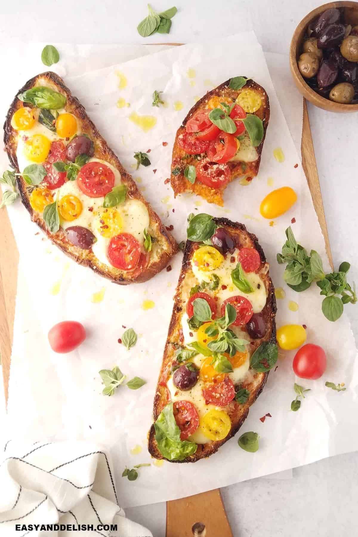 Pizza Toasts topped with mozzarella, colorful tomatoes, olives, and fresh herbs on a cutting board.