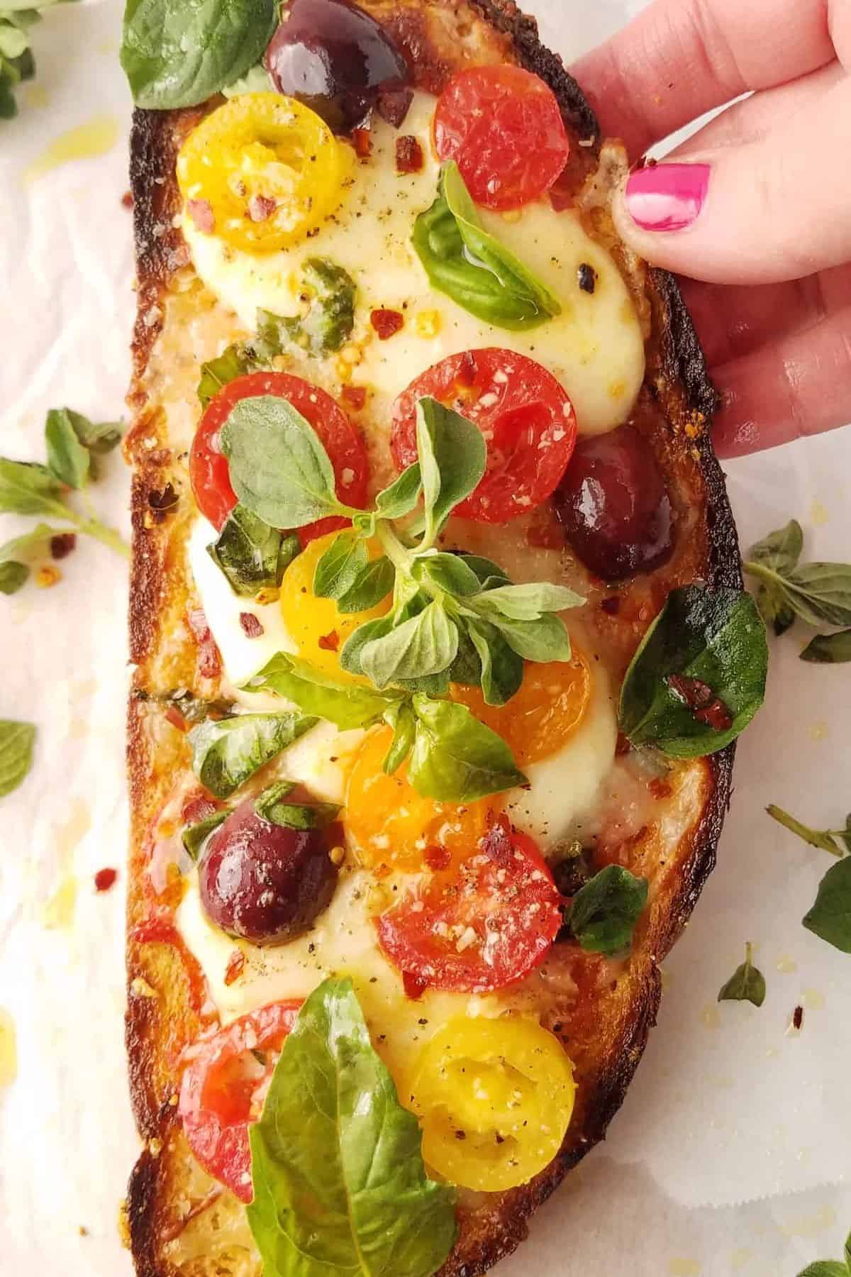 Hand holding a crisp toast topped with melted mozzarella, colorful tomatoes, olives, and fresh herbs.