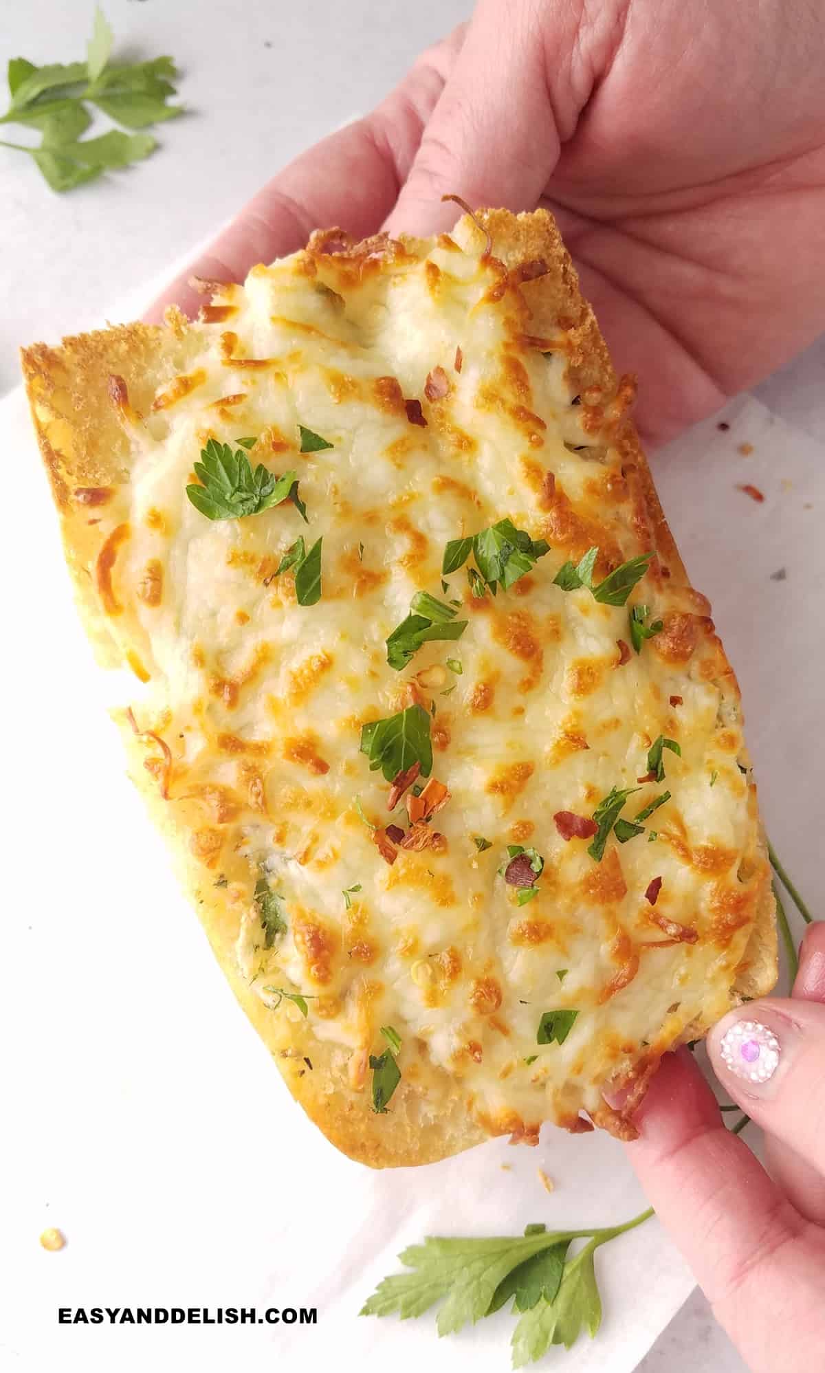 a hand holding a slice of cheesy baguette topped with chopped parsley.