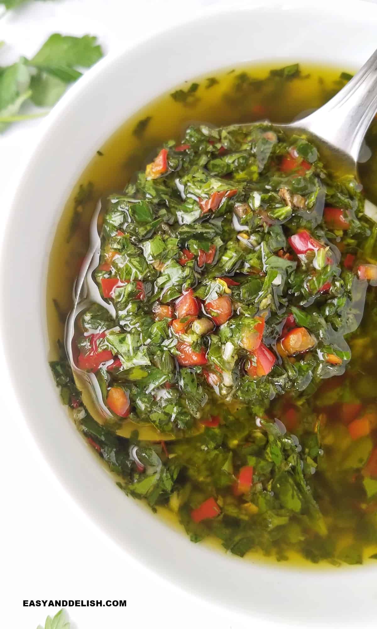 close up of a bowl of authentic chimichurri sauce with a spoon held over it.