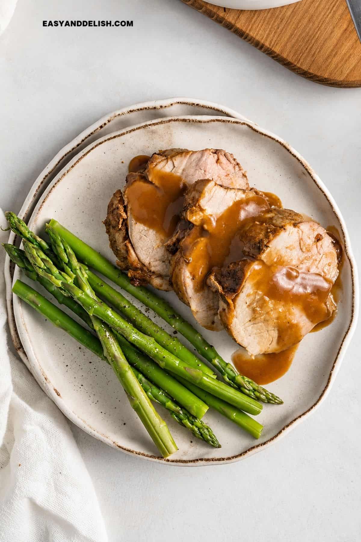 Slices of pork tenderloin topped with sauce beside cooked asparagus on a ceramic plate.