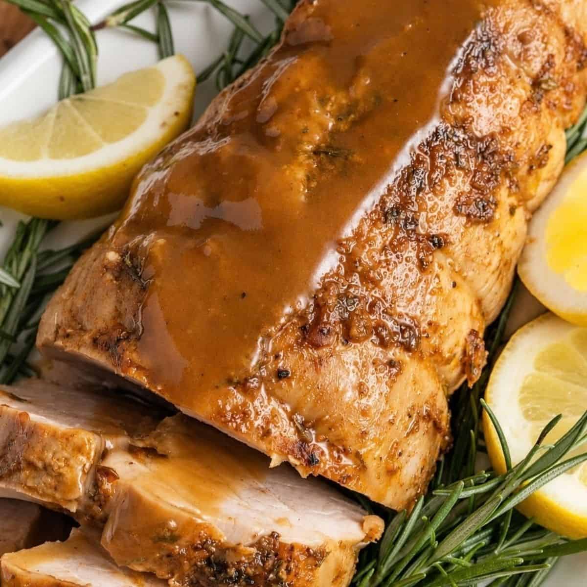 Sliced pork tenderloin topped with brown sauce and garnished with fresh rosemary and lemon wedges.