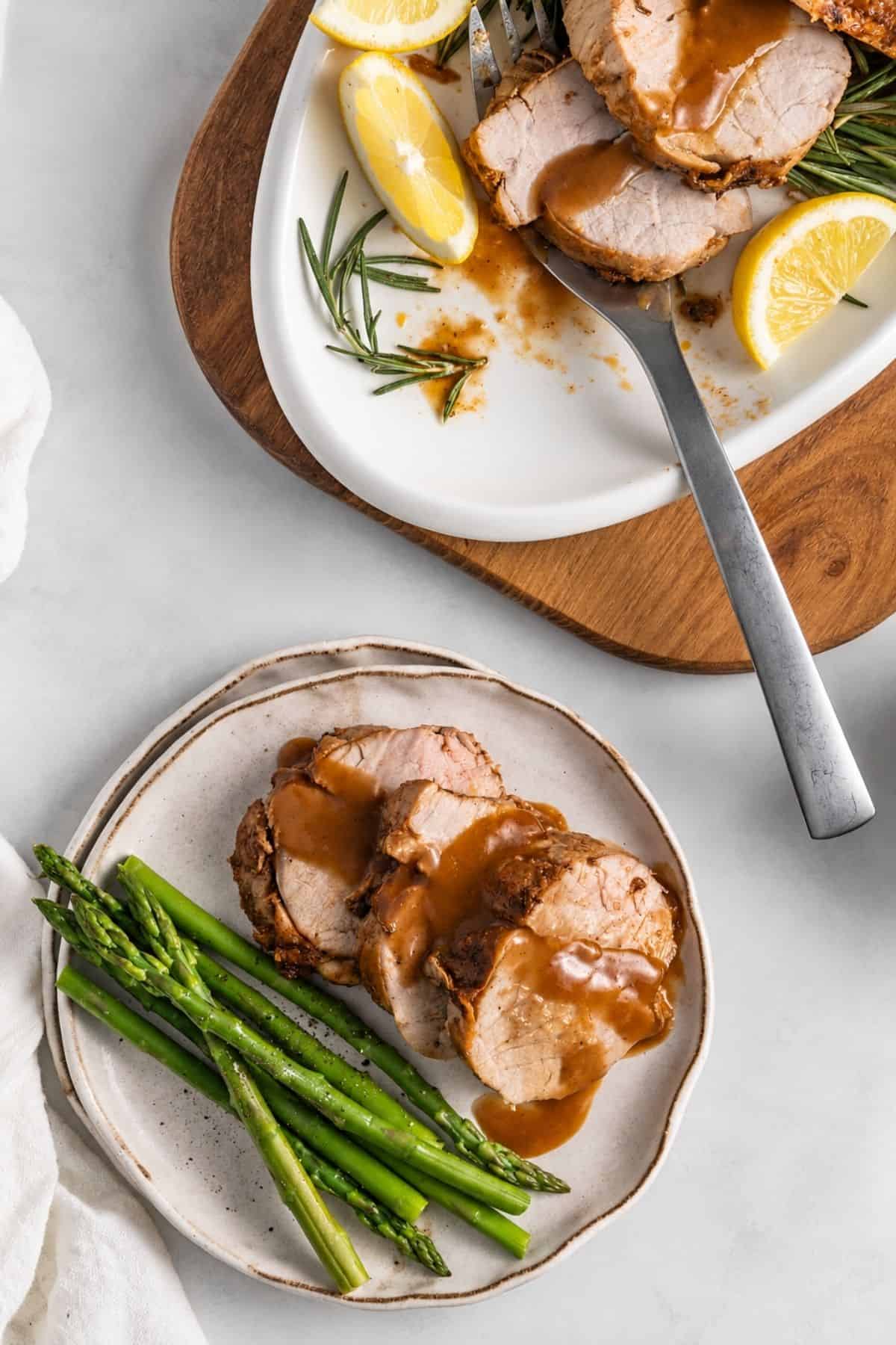 Sliced roasted pork with sauce, rosemary sprigs, and lemon wedges on a white platter, alongside a plate of pork and asparagus.