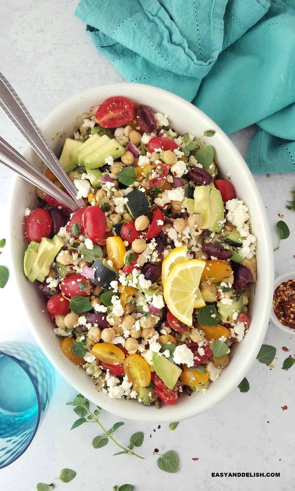 A bowl of cucumber chickpea salad with serving spoons plus some garnishes.