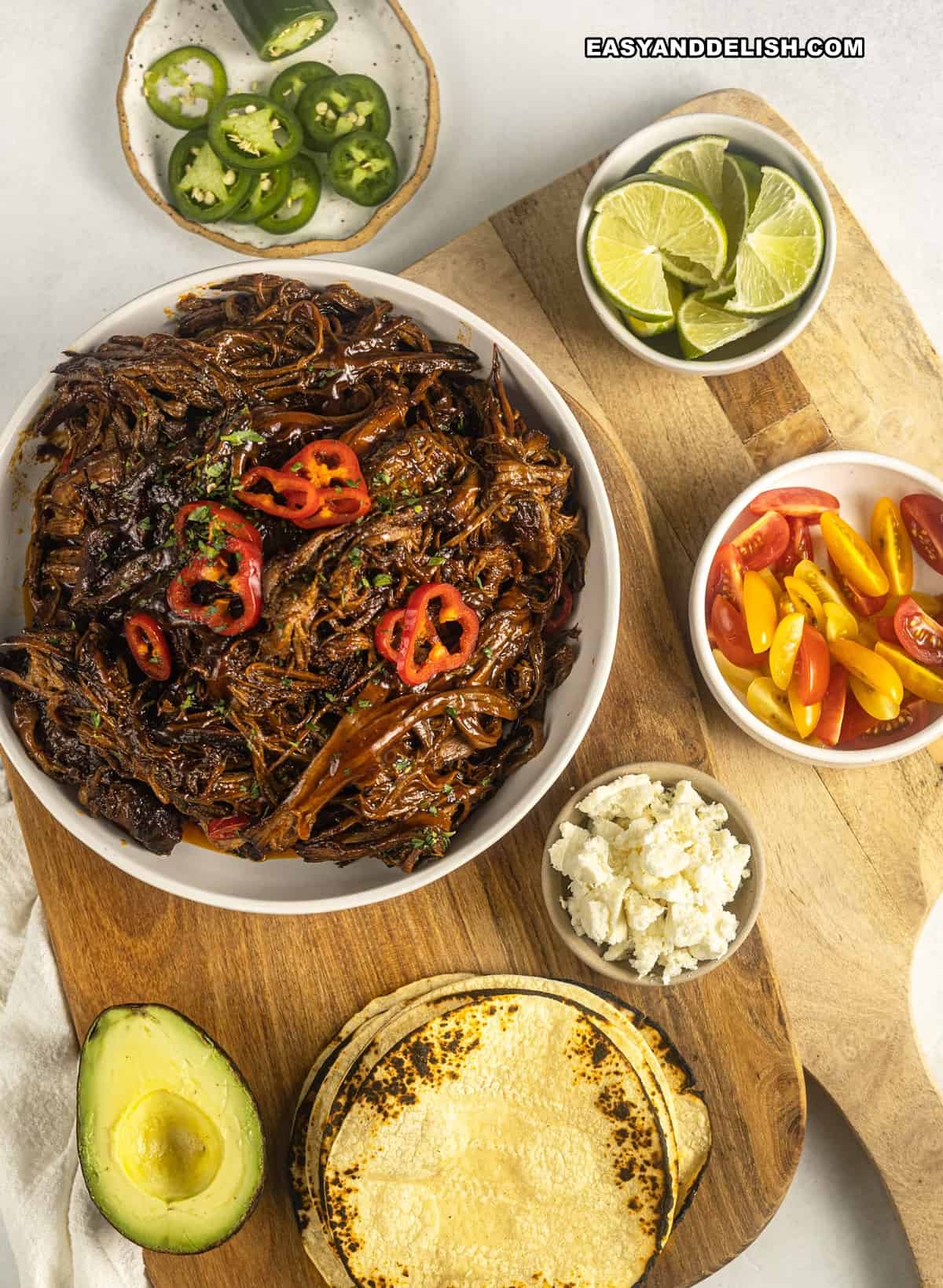 BBQ pulled beef in a bowl with garnishes and tortillas on the side.