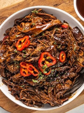 BBQ pulled brisket topped with sliced red peppers and fresh herbs in a white serving bowl.