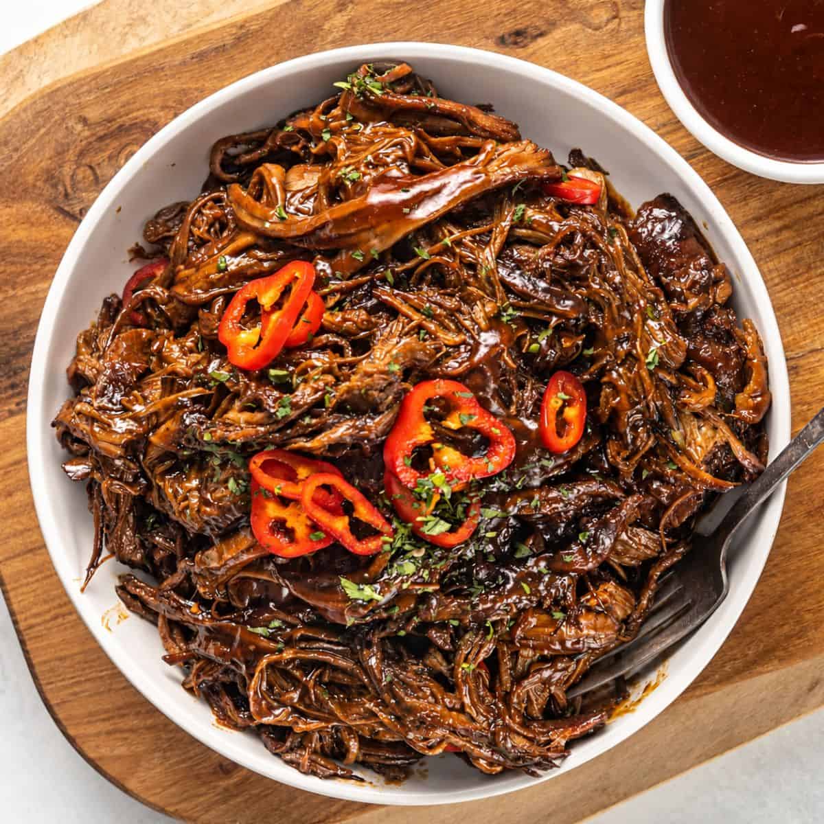 BBQ pulled brisket topped with sliced red peppers and fresh herbs in a white serving bowl.