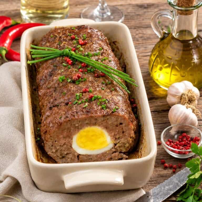 Baked meatloaf with a hard-boiled egg center and chive garnish in a rectangular ceramic dish.