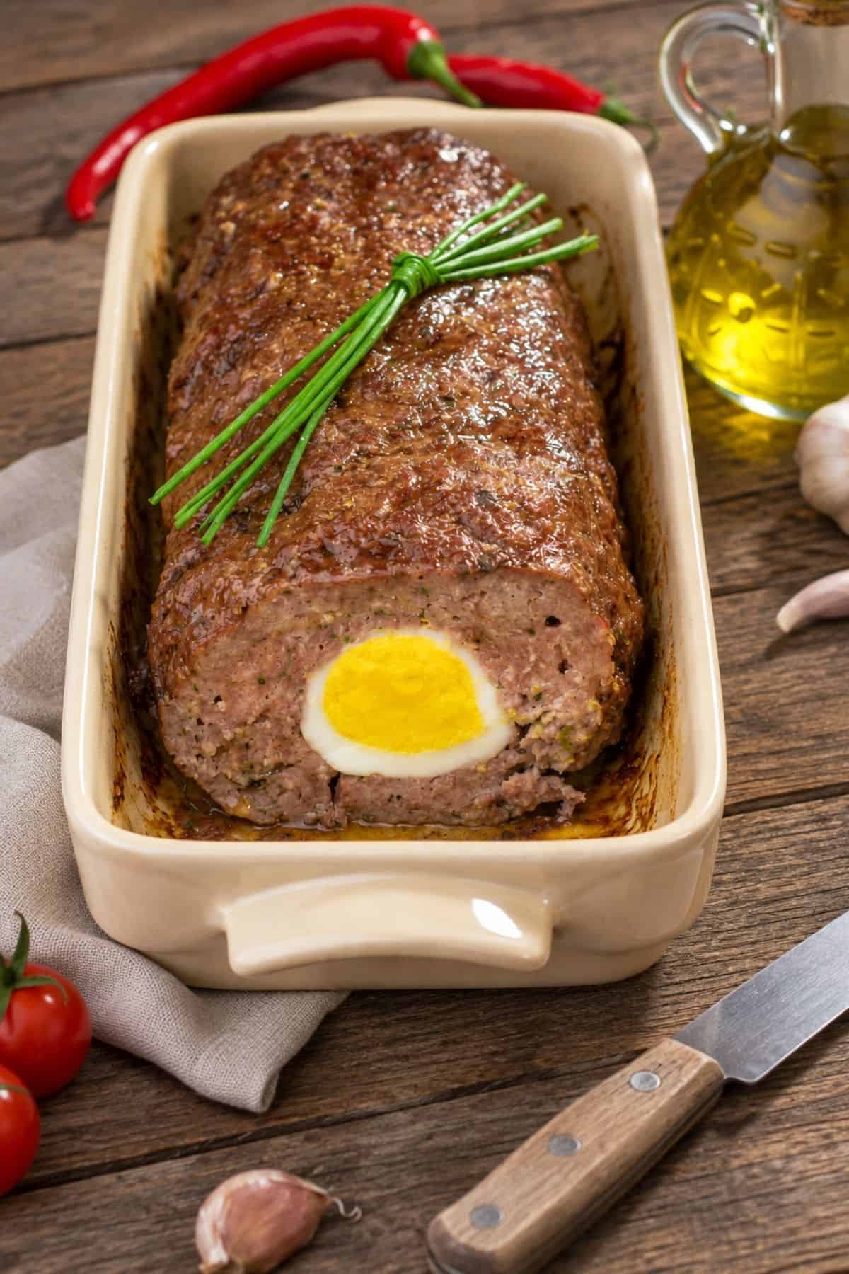 German Meatloaf in a green baking dish with a hard-boiled egg visible in the cut end and fresh chives on top.
