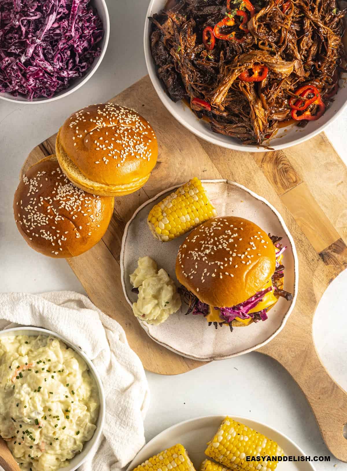 burger buns, bbq pulled brisket, and sides on table for assembling brisket sandwiches.