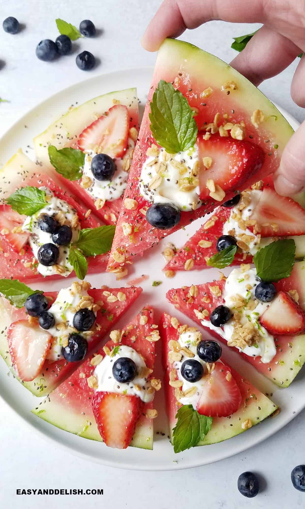 a hand holding a wedge of watermelon pizza with the rest of the treat under.