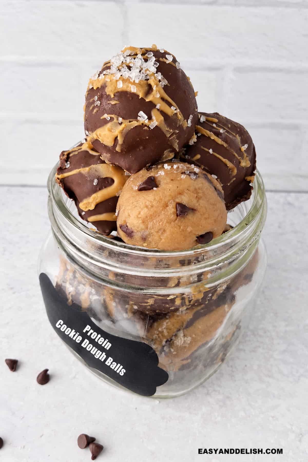 Plain and chocolate-coated protein balls stacked inside a glass jar, with chocolate chips scattered on the surface around it.