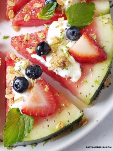 close up image of some slices of watermelon pizza topped with yogurt, berries, granola, honey, chia, and mint.