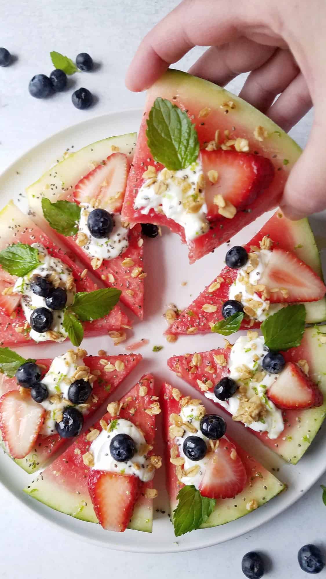 a hand holding a wedge of melon dessert pizza with yogurt and other toppings.