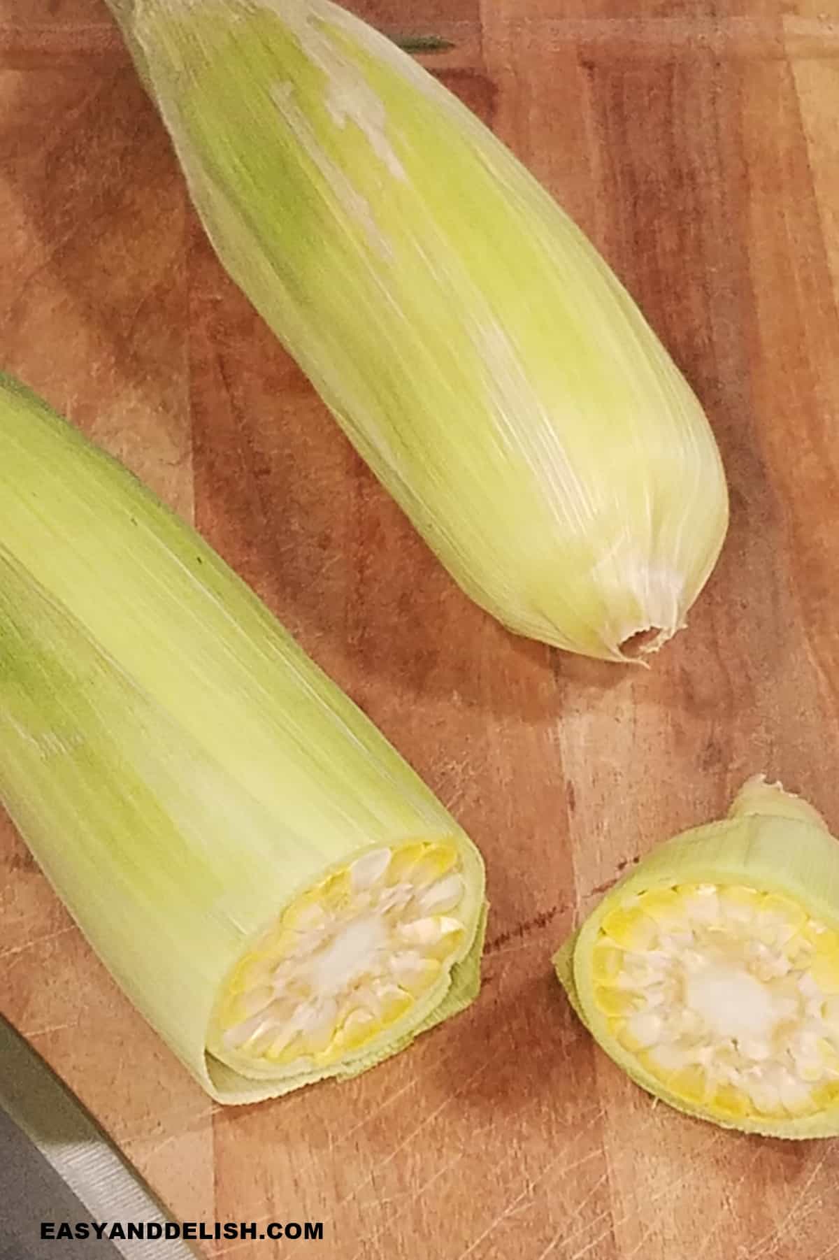 two ears of corn, one whole and another one with a cut-off end on a cutting board. 
