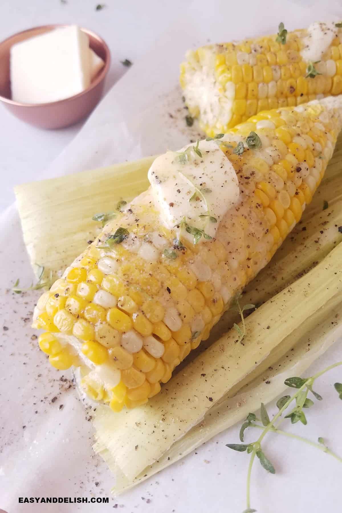 shucked corn on the cob over husk with some butter pat, seasonings,a nd herbs on top of the cob.
