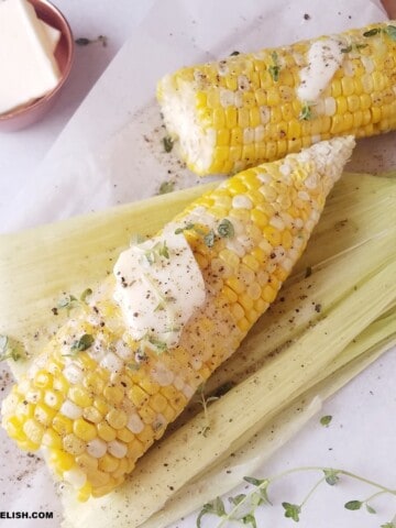 two cooked shucked corn on the cob with a at of butter and some seasonings on top.