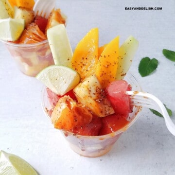 Mexican fruit cups on a table with fresh mint leaves and lime wedges around them.