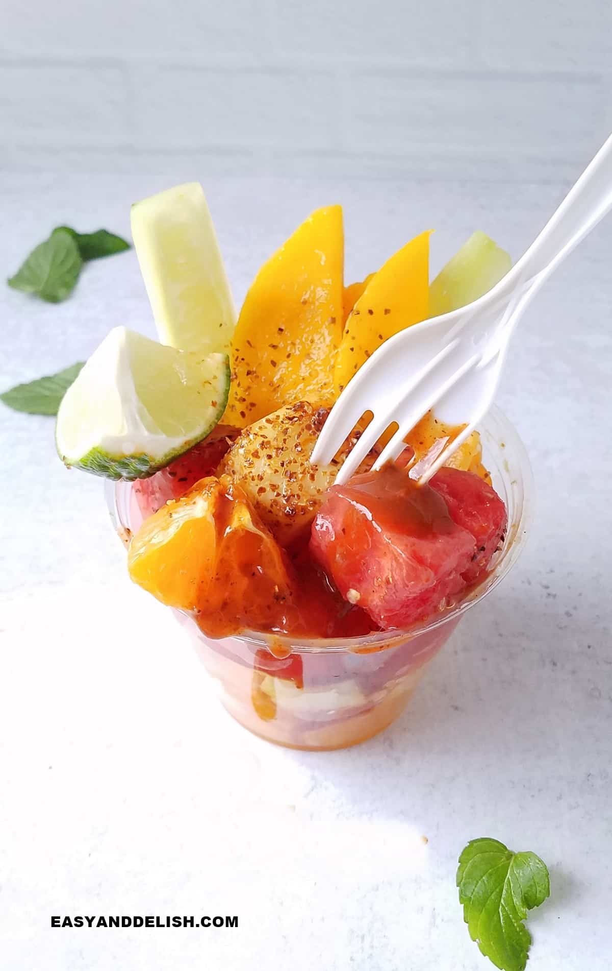 a mexican fruit cup with Chamoy and Tajin on a table with a fork picking a piece of fruit.