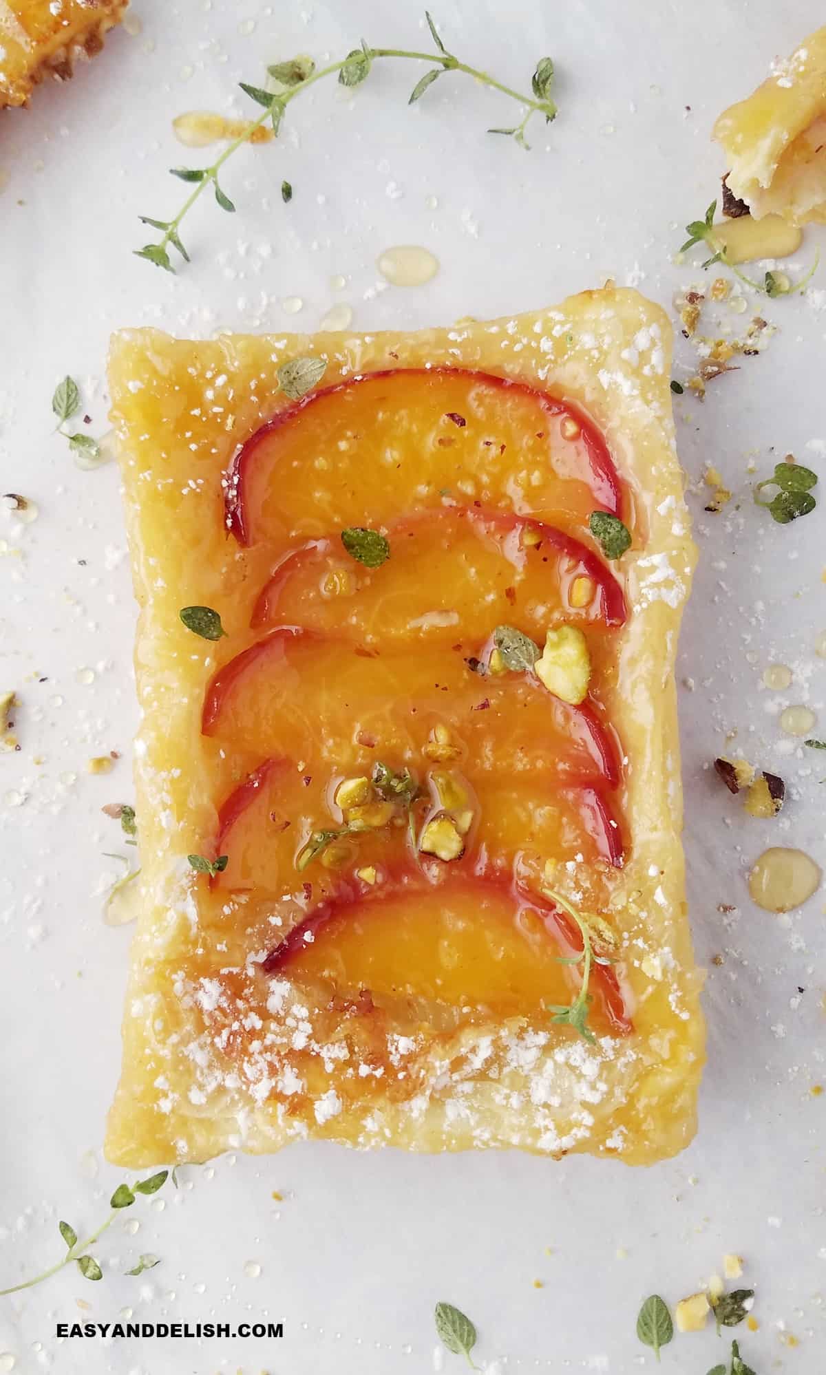 close up of a peach puff pastry tart oven parchment paper with herbs around it.
