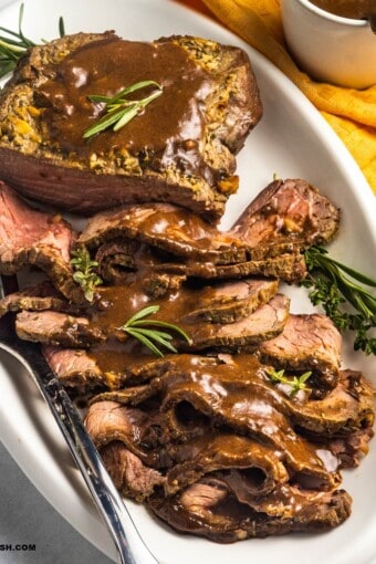 close up of a platter with sliced roast beef topped with bee gravy and garnished with fresh herbs.