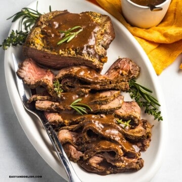 close up of a platter with sliced roast beef topped with bee gravy and garnished with fresh herbs.