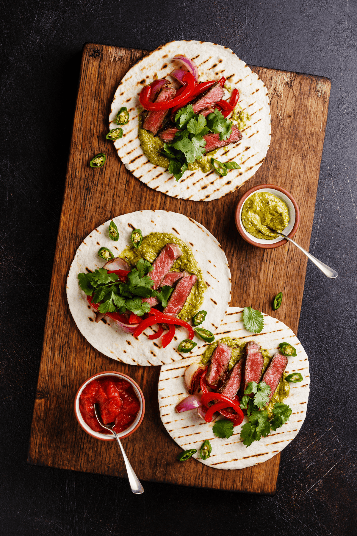 three tacos served over a cutting board with guacamole and salsa roja on the side.