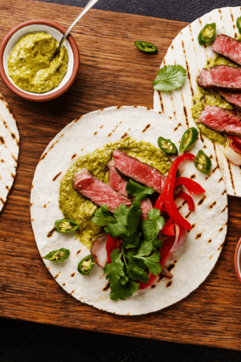 close up of flank steak tacos on a cutting board with some garnishes on the side.