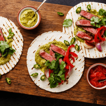 close up of flank steak tacos on a cutting board with some garnishes on the side.