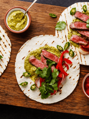 close up of flank steak tacos on a cutting board with some garnishes on the side.