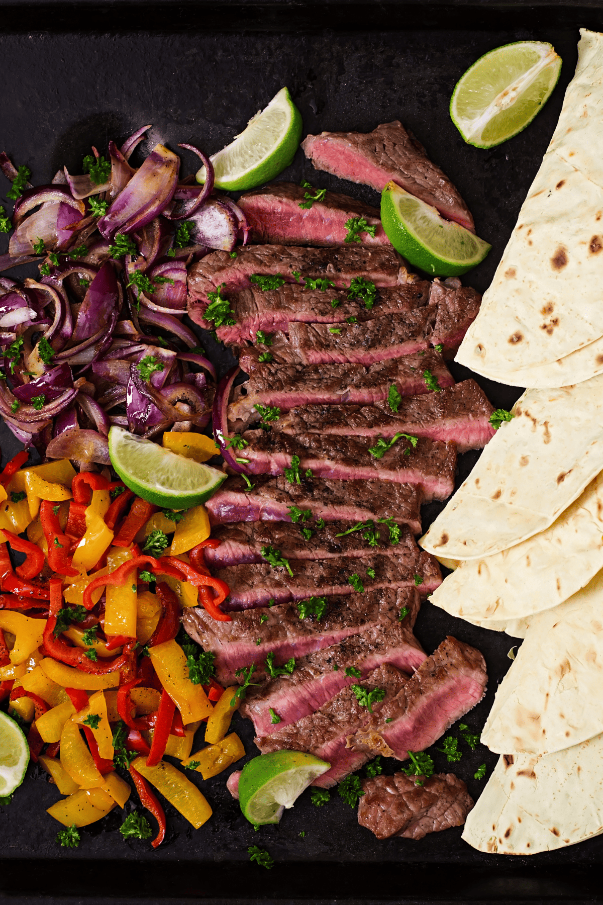 sliced grilled flank steak with tortillas, cooked veggies, and lime wedges in a tray.