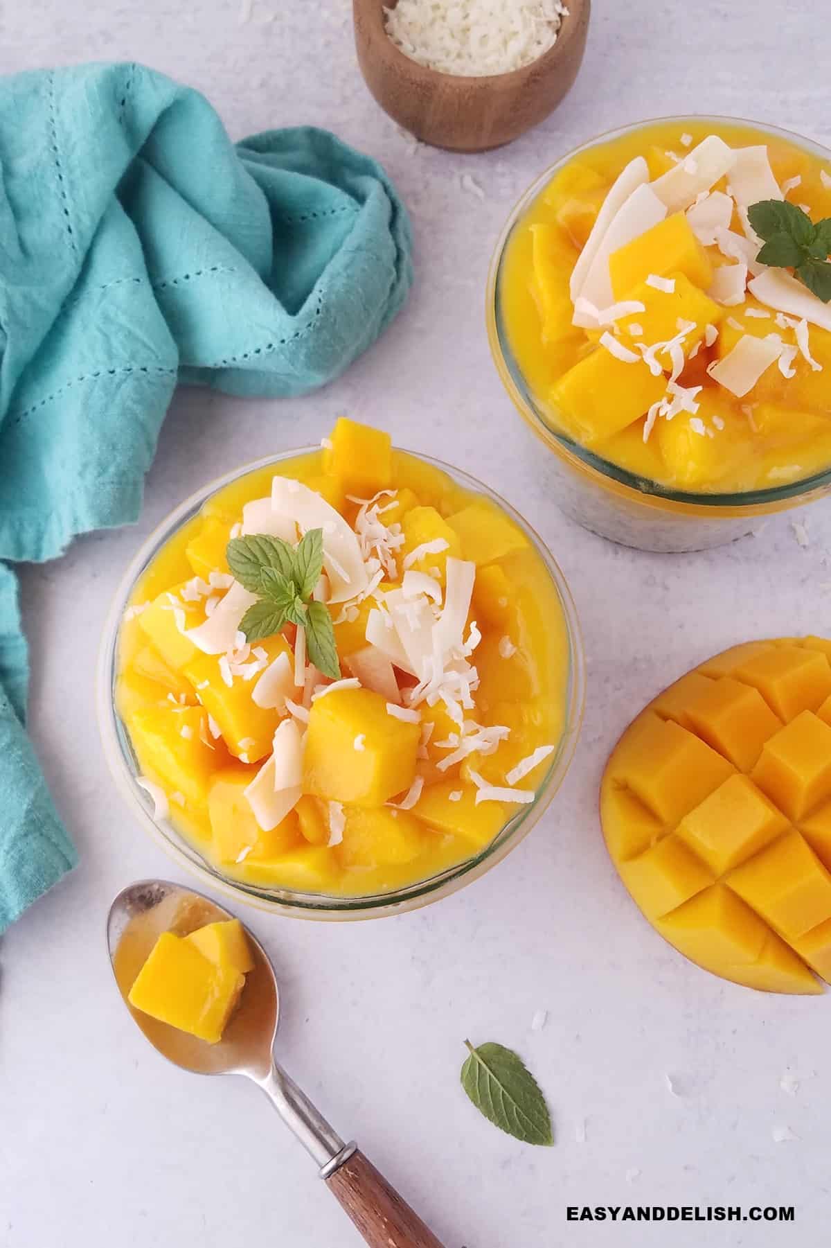 two mason jars of chia pudding with cconut milk and mango puree with mango chunks and coconut flakes plus a napkin, a spoon, and some garnishes.