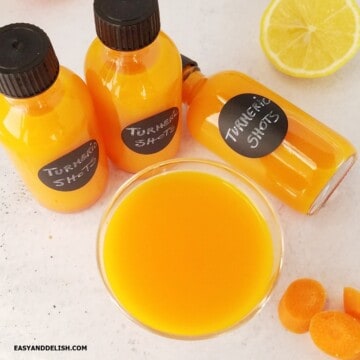 A shot glass and shot bottles containing turmeric shots and some ingredients around them.