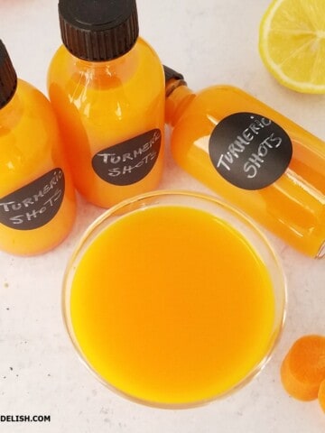 A shot glass and shot bottles containing turmeric shots and some ingredients around them.