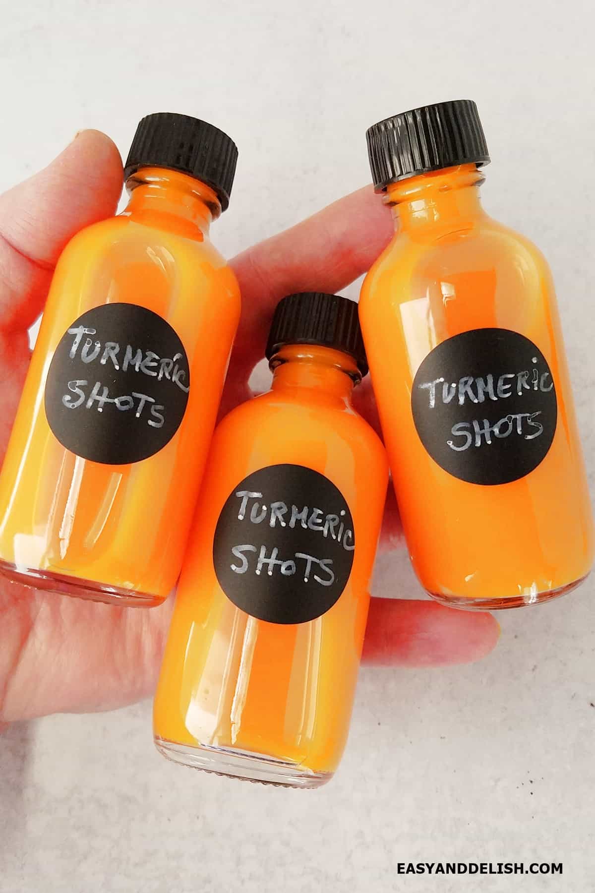 A hand holding flat three bottles of turmeric wellness shots.