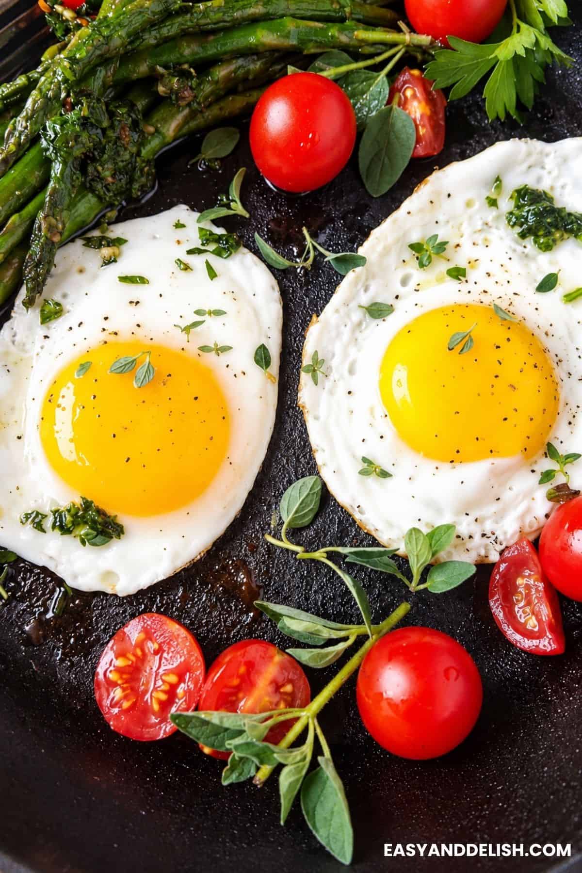 Two fried eggs in a skillet with cherry tomatoes, asparagus, and fresh herbs.