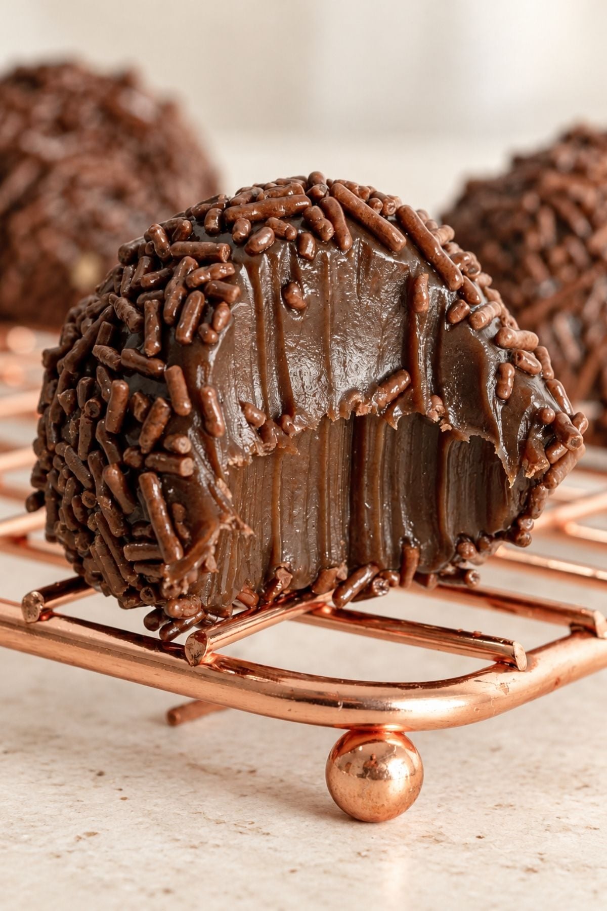 Close-up of a cut-open chocolate brigadeiro with a glossy, fudgy center and chocolate sprinkles, resting on a copper-colored wire rack.
