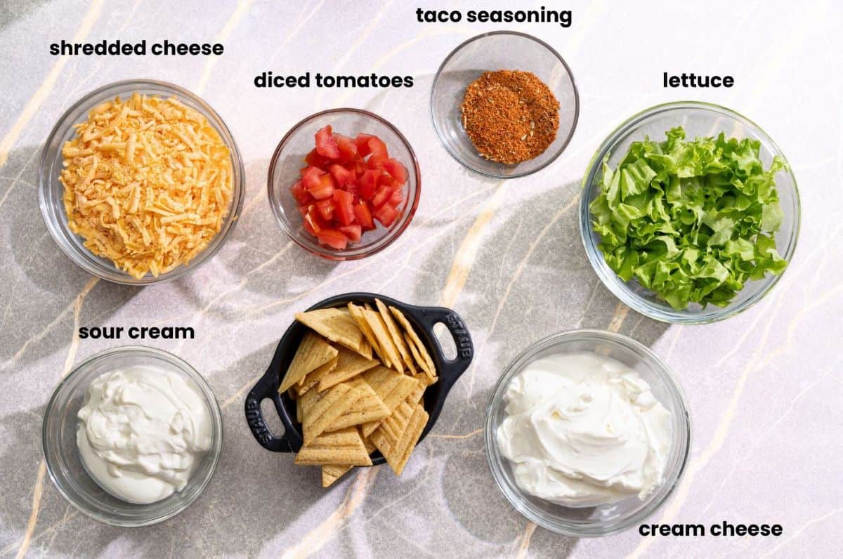 ingredients for easy taco dip recipe including cream cheese sour cream lettuce and cheddar cheese.