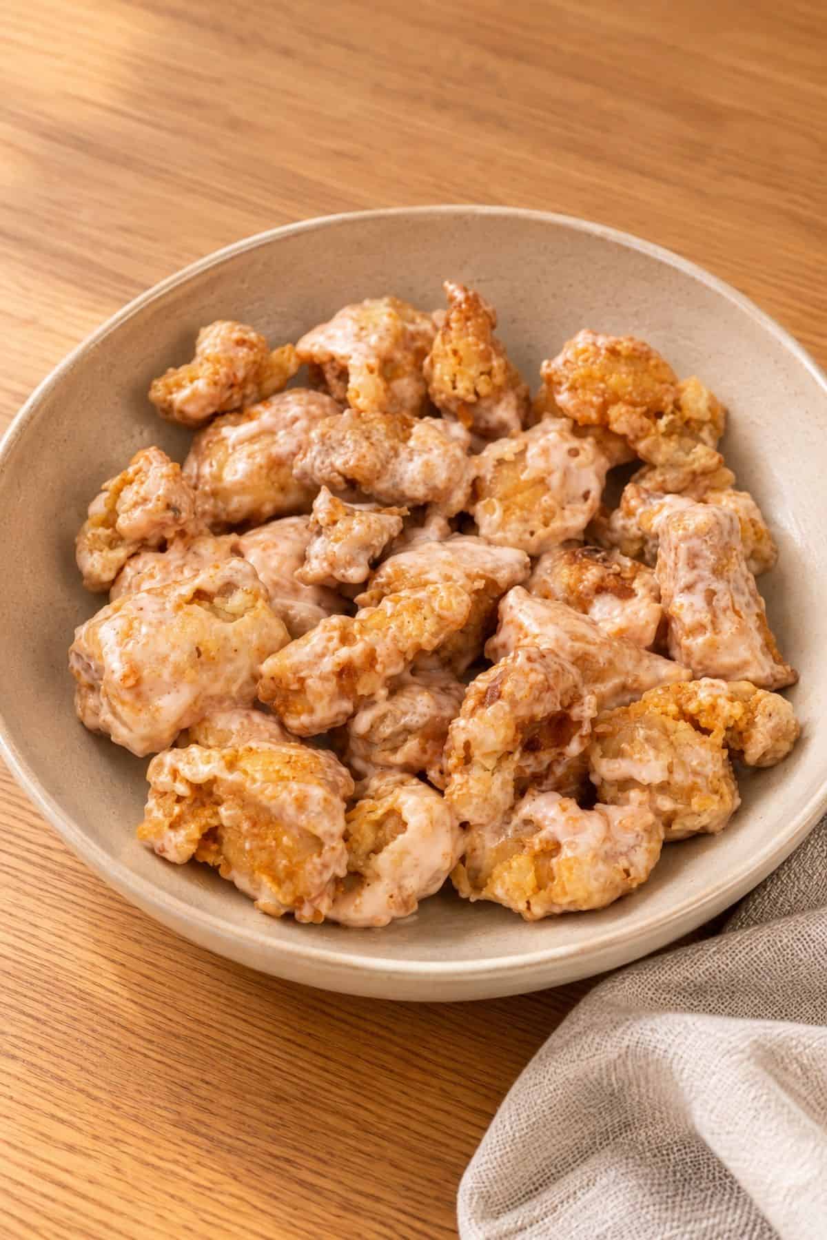 Crispy Bang Bang Chicken coated in a sauce in a beige ceramic bowl on a wooden table.